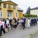 Grupa ludzi w eleganckich strojach idzie ulicą obok żółtego budynku. W tle zieleń i kolejny dom. Osoby niosą chleb na tacach.