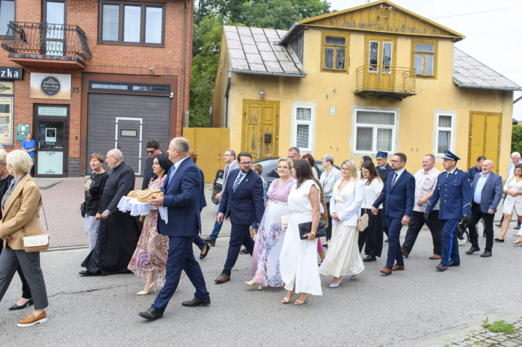 Grupa elegancko ubranych osób idzie ulicą. W tle ceglana kamienica i żółty dom. Atmosfera uroczystości formalnej.