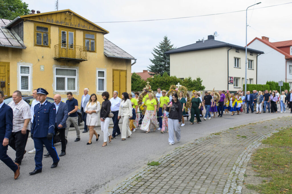 Grupa ludzi idzie ulicą obok żółtego domu z balkonem. Uczestnicy ubrani są kolorowo, w tle widać drzewa i budynki. Chodnik jest brukowany, a ludzie uczestniczą w czymś w rodzaju procesji.