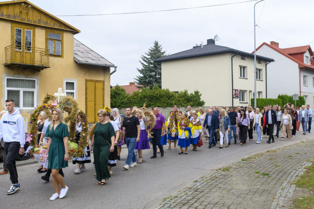 Grupa ludzi idzie ulicą podczas procesji dożynkowej. Ubrania w kolorach zieleni i fioletu, a tło to żółty budynek i krzewy. Widać ozdobne wieńce z kwiatów i zbóż.