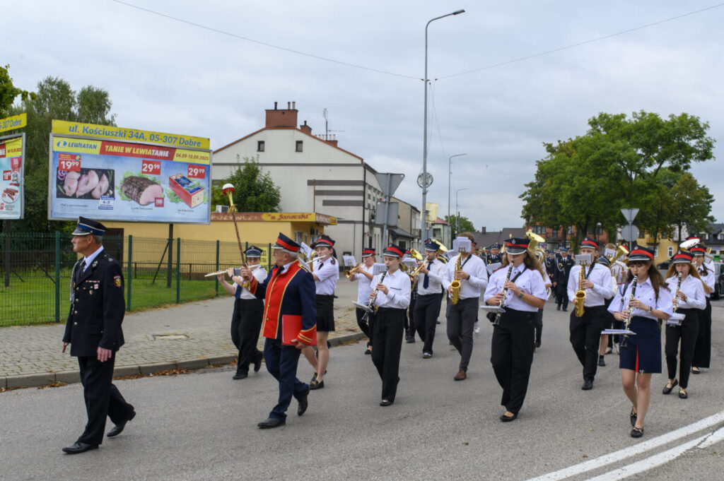 Parada orkiestry dętej w białych koszulach i czarnych spodniach, maszerującej ulicą. W tle zielone drzewa i reklamy supermarketu. Uczestnicy grają na instrumentach dętych i perkusyjnych.