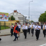 Parada orkiestry dętej w białych koszulach i czarnych spodniach, maszerującej ulicą. W tle zielone drzewa i reklamy supermarketu. Uczestnicy grają na instrumentach dętych i perkusyjnych.