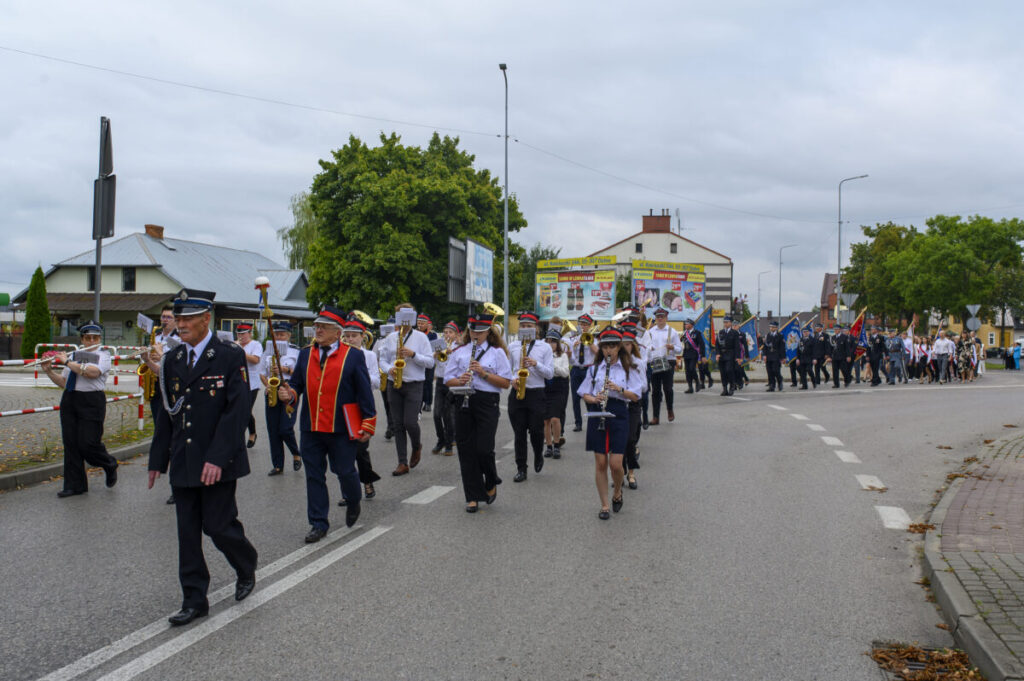 Orkiestra dęta maszeruje ulicą, ubrani w białe koszule i czarne spodnie pod pochmurnym niebem. Prowadzi osoba w granatowym mundurze z czerwonymi akcentami. W tle znajdują się drzewa i budynki z kolorowymi reklam