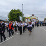 Orkiestra dęta maszeruje ulicą, ubrani w białe koszule i czarne spodnie pod pochmurnym niebem. Prowadzi osoba w granatowym mundurze z czerwonymi akcentami. W tle znajdują się drzewa i budynki z kolorowymi reklam