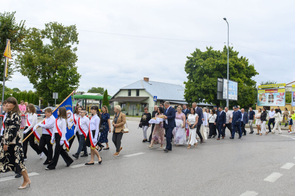 Parada ludzi na ulicy; dzieci w białych koszulach z flagami. Tło to drzewa i budynki. Ciepły letni dzień.