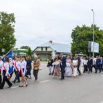 Parada ludzi na ulicy; dzieci w białych koszulach z flagami. Tło to drzewa i budynki. Ciepły letni dzień.