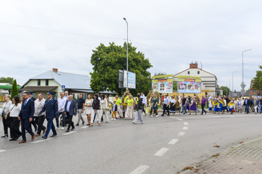 Grupa ludzi maszeruje ulicą, ubrani w różne kolory. W tle zielone drzewo i budynek z reklamami. Niebo jest zachmurzone.