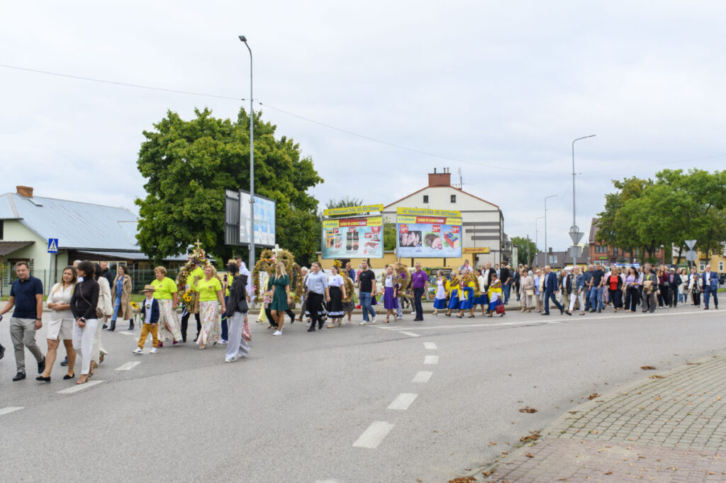Grupa ludzi idzie w korowodzie ulicą. Uczestnicy noszą kolorowe stroje ludowe i niosą wieńce. W tle są budynki i drzewa.