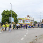 Grupa ludzi idzie w korowodzie ulicą. Uczestnicy noszą kolorowe stroje ludowe i niosą wieńce. W tle są budynki i drzewa.