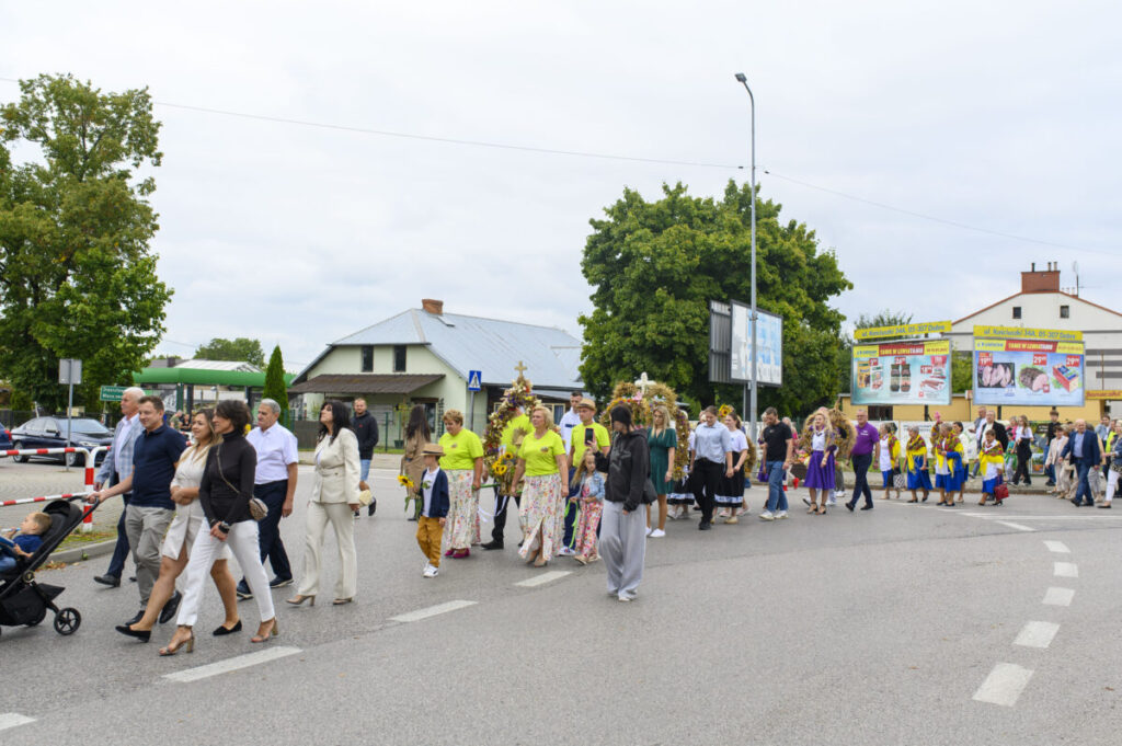 Grupa ludzi maszeruje ulicą podczas procesji, wielu ubranych kolorowo. W tle zielone drzewa i reklamy. Atmosfera jest świąteczna.
