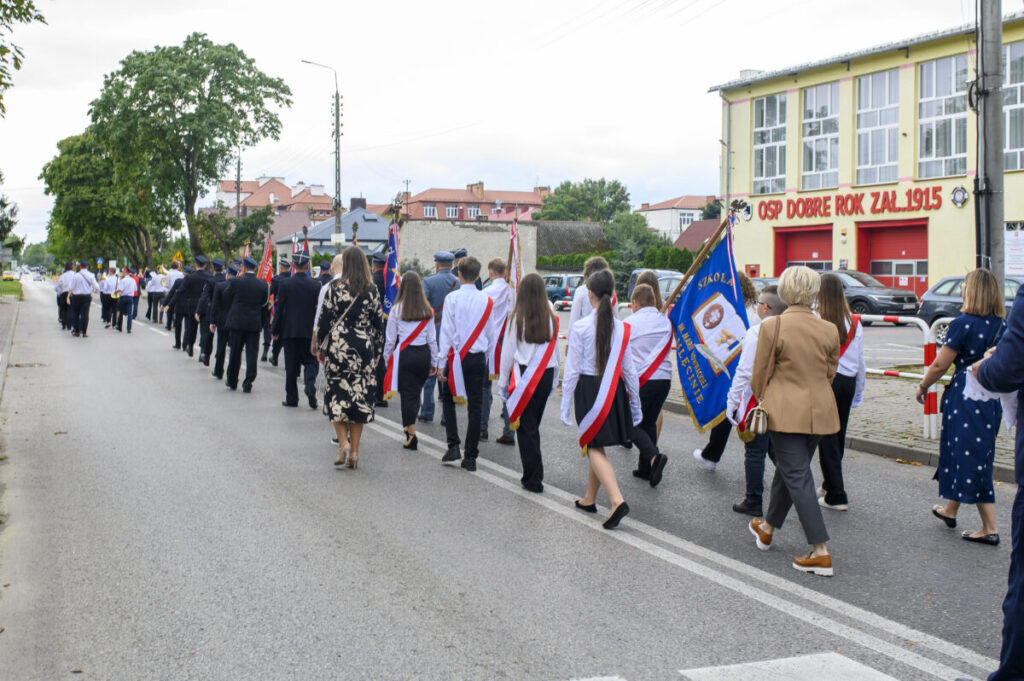 Grupa osób z flagami idzie ulicą w pobliżu remizy strażackiej. Dominują kolory biały, czerwony i niebieski. W tle są drzewa i budynki mieszkalne.