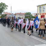 Grupa osób z flagami idzie ulicą w pobliżu remizy strażackiej. Dominują kolory biały, czerwony i niebieski. W tle są drzewa i budynki mieszkalne.