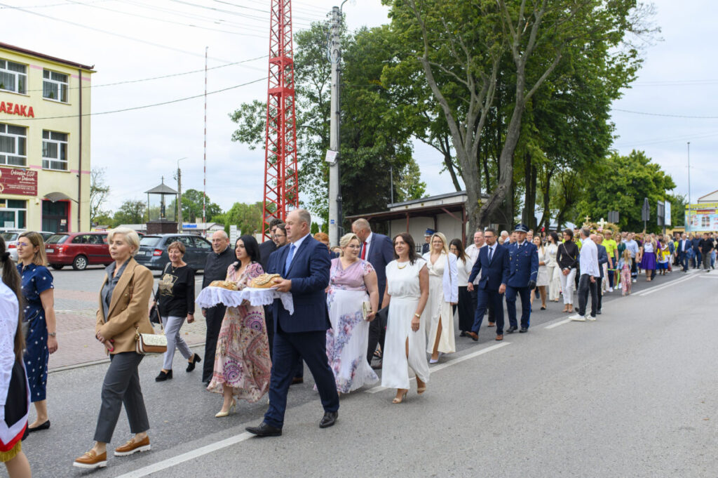 Grupa elegancko ubranych ludzi maszeruje ulicą. Niebo jest pochmurne, a w tle widoczne są drzewa i budynki. Na czołowym planie osoba niesie chleb na białej serwetce.