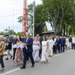 Grupa elegancko ubranych ludzi maszeruje ulicą. Niebo jest pochmurne, a w tle widoczne są drzewa i budynki. Na czołowym planie osoba niesie chleb na białej serwetce.
