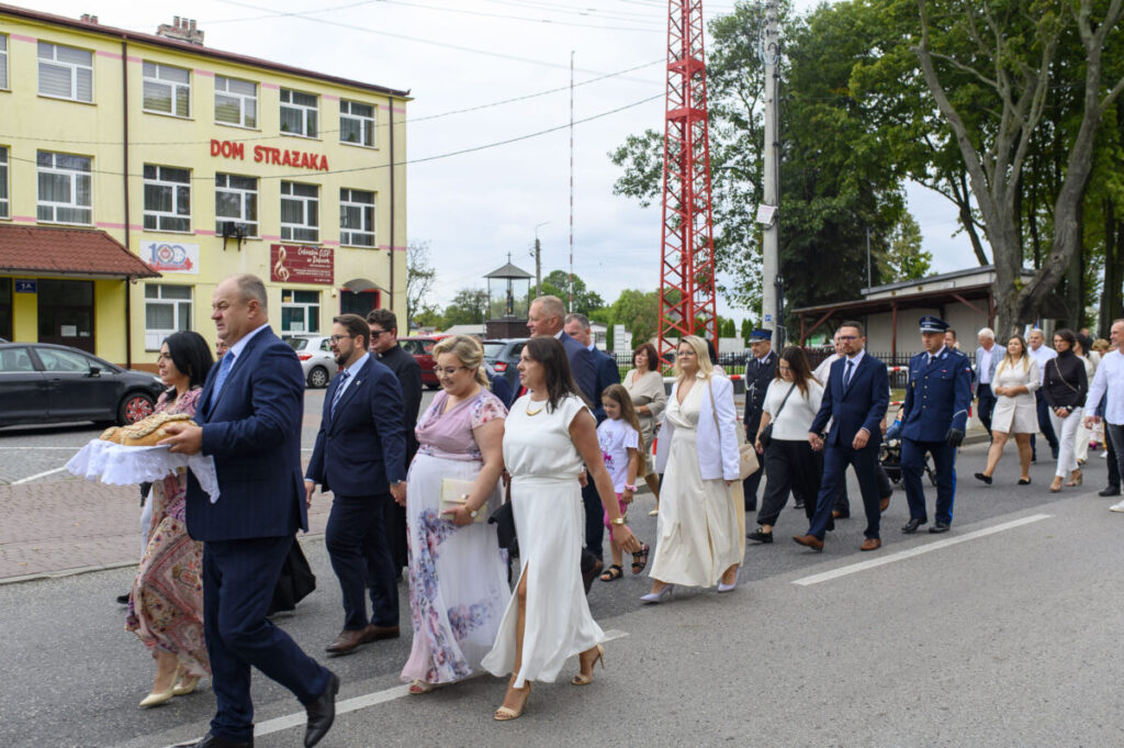 Grupa elegancko ubranych ludzi idzie ulicą. W tle żółty budynek z napisem Dom Strażaka. Po prawej wysokie drzewa i wieża.