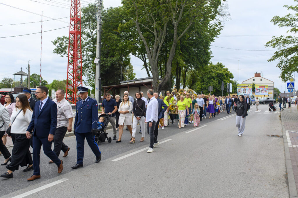Grupa osób w różnokolorowych ubraniach maszeruje po ulicy. W tle drzewa i budynki. Pogoda pochmurna.