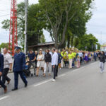 Grupa osób w różnokolorowych ubraniach maszeruje po ulicy. W tle drzewa i budynki. Pogoda pochmurna.