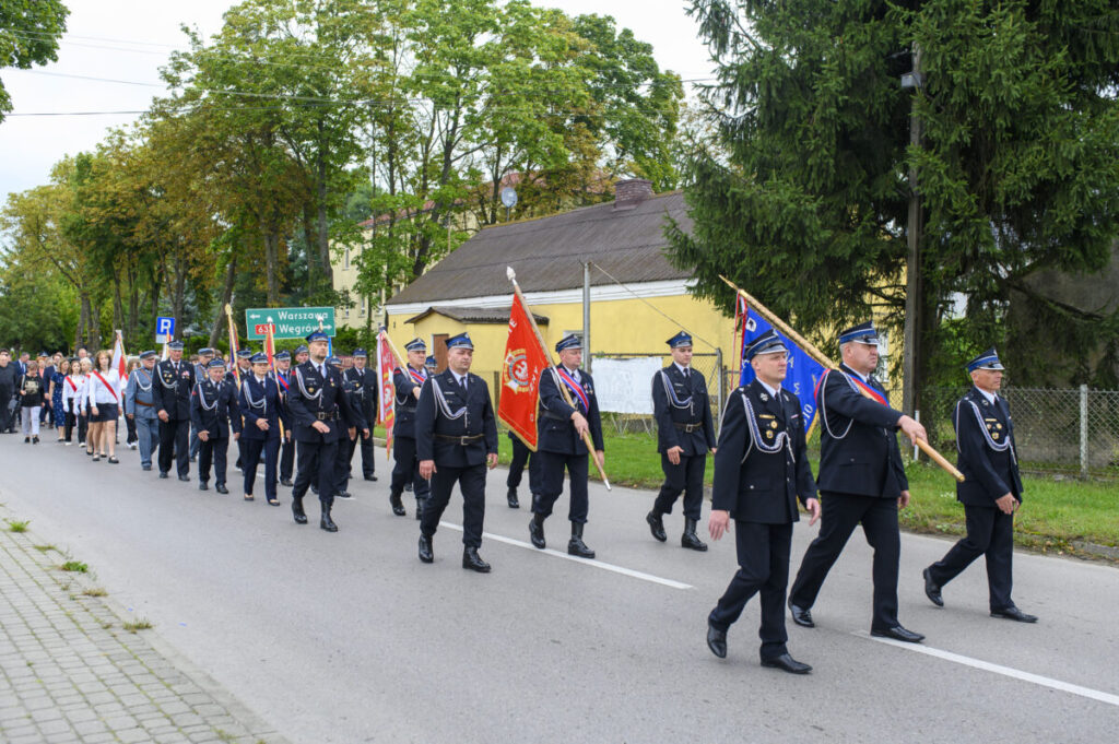 Parada strażaków w mundurach na ulicy, niosących flagi. Tło wypełnia zielona roślinność i budynki. Niebo zachmurzone.