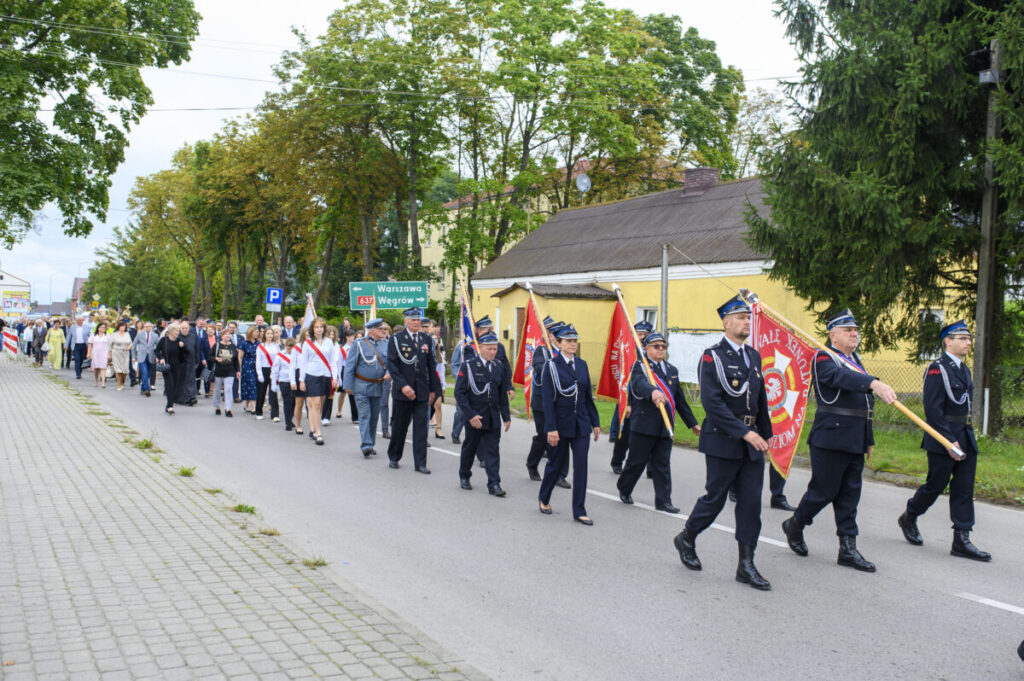Parada strażaków i mieszkańców w mundurach na ulicy. Zielone drzewa, żółty budynek w tle. Flagi czerwone i niebieskie.