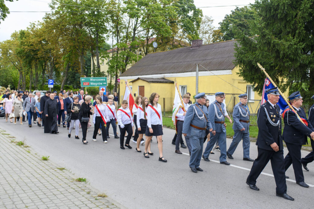 Grupa ludzi w paradzie na ulicy, niektórzy w mundurach z flagami. W tle żółty budynek i zielona roślinność. Długa procesja z tablicą informacyjną.
