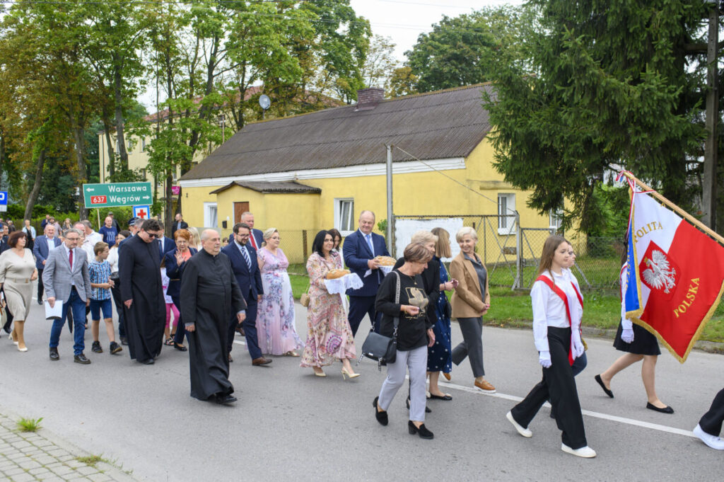 Grupa ludzi maszeruje ulicą obok żółtego budynku, niesie flagę Polski. W tle drzewa i zielony znak drogowy. Atmosfera uroczysta.