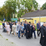 Grupa ludzi idzie ulicą, otoczona zielonymi drzewami i żółtymi budynkami. Uczestnicy noszą eleganckie stroje. W tle widoczne są znaki drogowe.