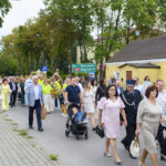 Grupa ludzi w różnorodnych strojach maszeruje ulicą. Widoczne drzewa i żółty budynek w tle. Wózek z dzieckiem na czele.