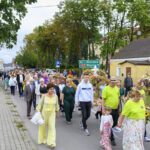 Na zdjęciu widać tłum ludzi maszerujących ulicą w jasnych kolorach. W tle są drzewa i żółty budynek. Uczestnicy niosą barwne wieńce, a nad głowami widać znak drogowy.