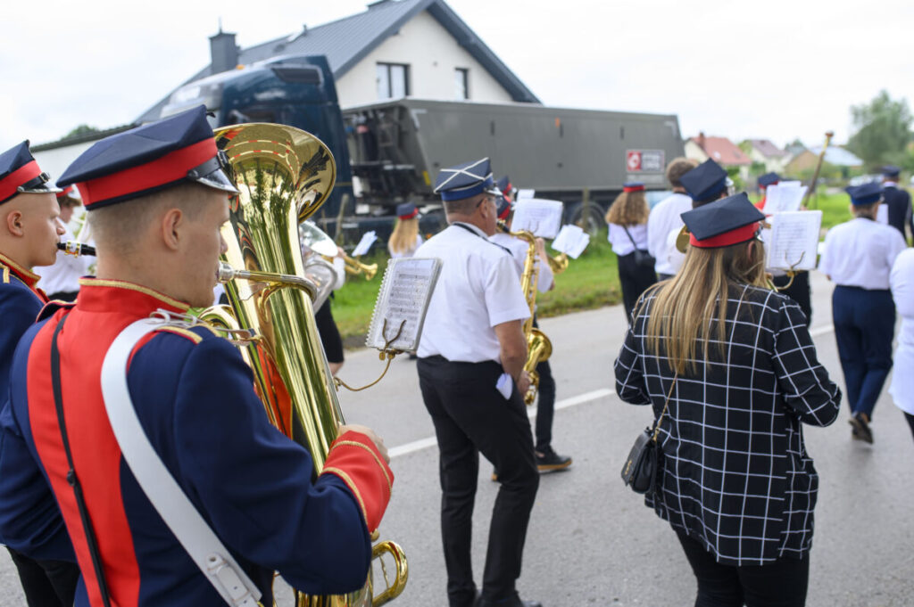 Grupa muzyków maszeruje ulicą w strojach paradnych. W tle widać budynek i ciężarówkę. Dominują kolory niebieski, biel i czerwień.