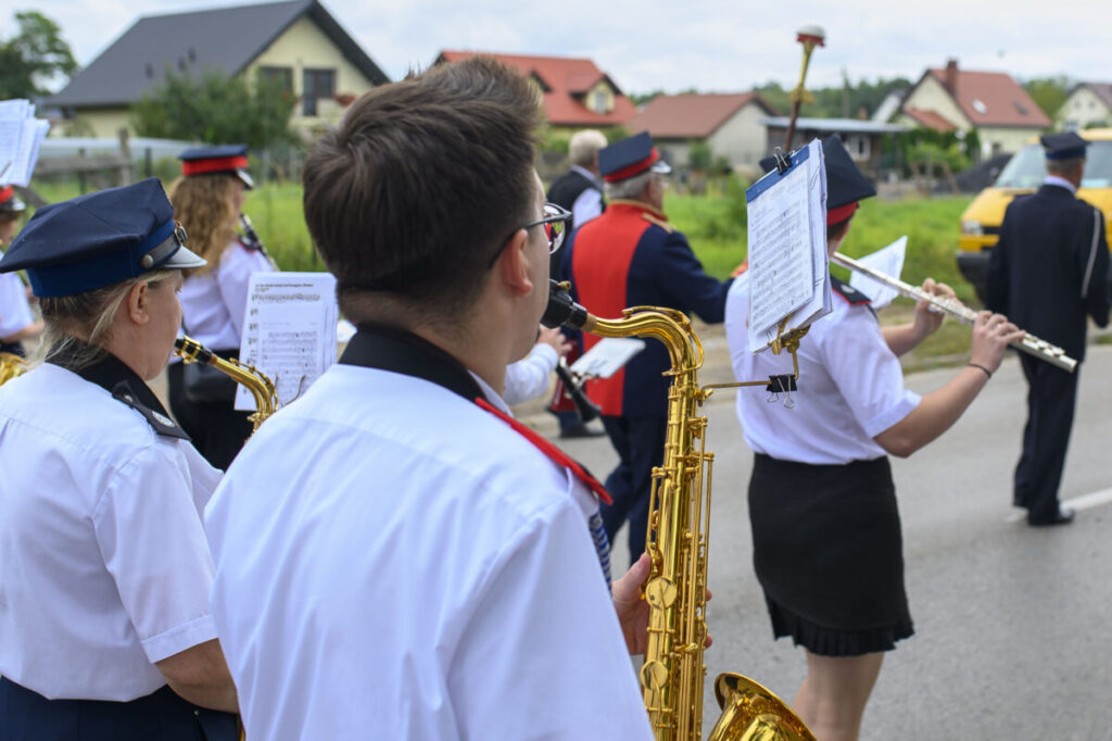 Orkiestra dęta na ulicy. Członkowie w białych koszulach grają na instrumentach. W tle domy i zielone tło.