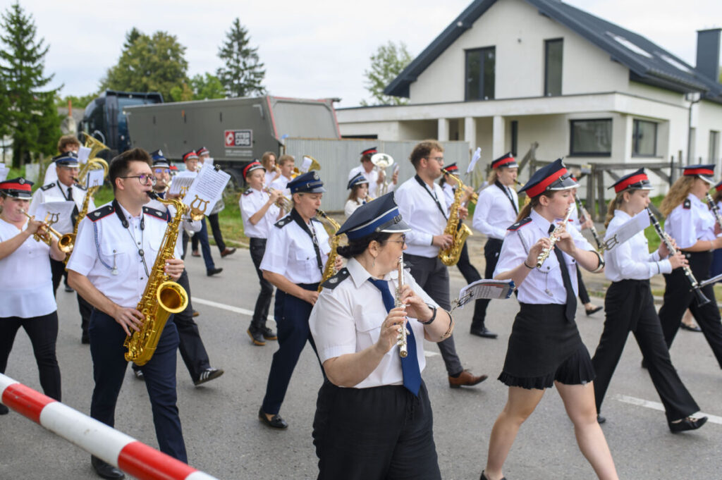 Orkiestra dęta w białych koszulach i czapkach maszeruje ulicą. Muzycy grają na instrumentach w złotym kolorze. Za nimi w tle dom i drzewa.