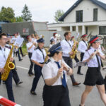 Orkiestra dęta w białych koszulach i czapkach maszeruje ulicą. Muzycy grają na instrumentach w złotym kolorze. Za nimi w tle dom i drzewa.