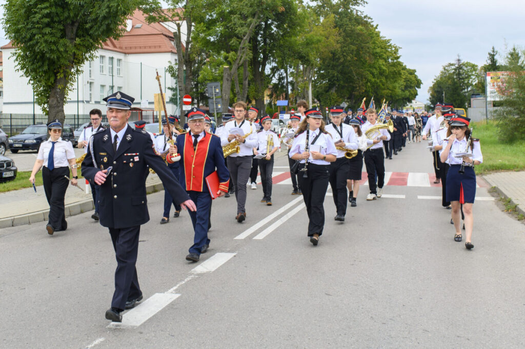 Orkiestra maszeruje ulicą w strojach galowych, grając instrumenty dęte. Tło to drzewa i budynek z czerwoną dachówką. Pogoda jest pochmurna.