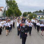 Orkiestra maszeruje ulicą, członkowie w białych koszulach i czapkach. W tle drzewa i budynki. Niebo zachmurzone.