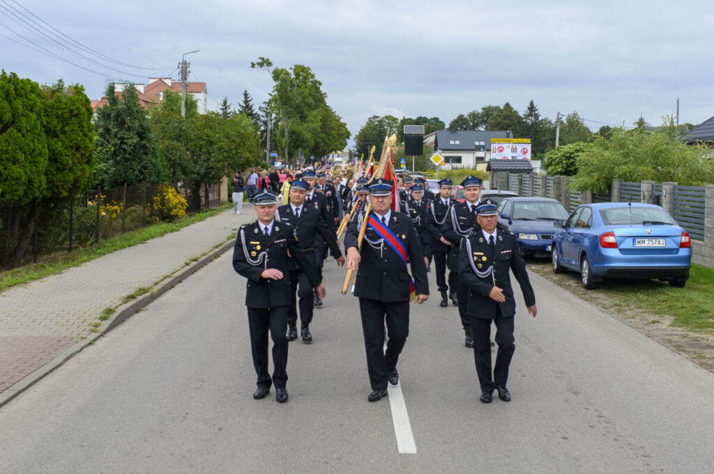 Strażacy w czarnych mundurach maszerują po ulicy. Niosą sztandary, a tło to zieleń i budynki. Obok parkują auta.