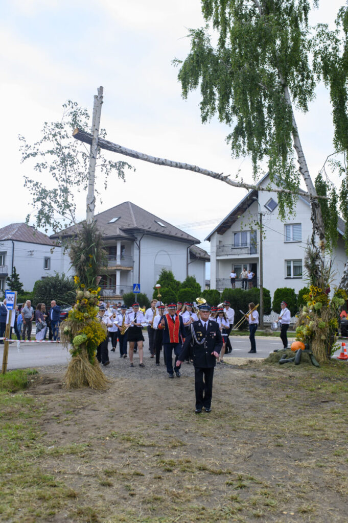 Parada orkiestry dętej w mundurach maszeruje drogą. Otoczenie to domy i drzewa, na trawie leżą dynie. Pogoda pochmurna.