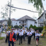 Orkiestra dęta w mundurach maszeruje przez wieś. Dominują kolory czerwony i niebieski, jest pochmurnie. W tle są domy i drzewa.