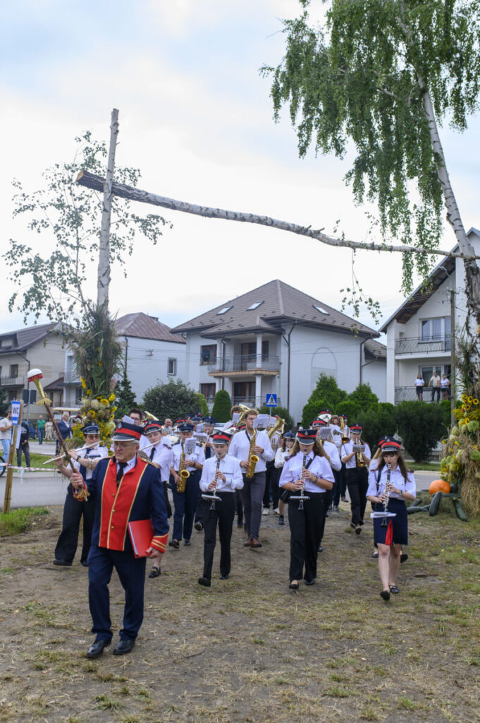 Orkiestra dęta w mundurach maszeruje przez wieś. Dominują kolory czerwony i niebieski, jest pochmurnie. W tle są domy i drzewa.