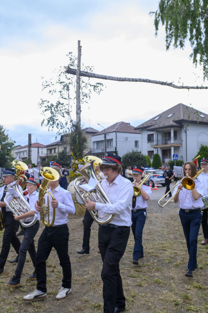 Orkiestra dęta w białych koszulach idzie po trawie, trzymając instrumenty. W tle domy i błękitne niebo.