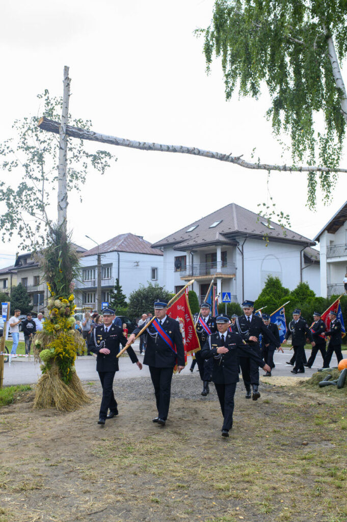 Grupa strażaków maszeruje z czerwonymi flagami, ubrani w ciemne mundury. Tło stanowią domy i zielone drzewa. Nad nimi drewniana konstrukcja z dekoracjami z kwiatów i traw.