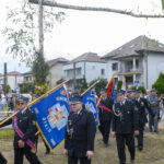 Strażacy w mundurach maszerują z niebieskimi sztandarami. W tle domy i zielone drzewa. Uroczystość odbywa się na zewnątrz.