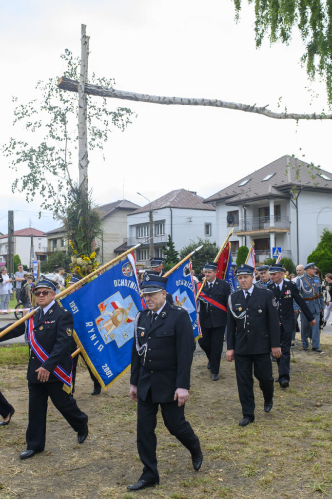 Strażacy w mundurach maszerują z niebieskimi sztandarami. W tle domy i zielone drzewa. Uroczystość odbywa się na zewnątrz.