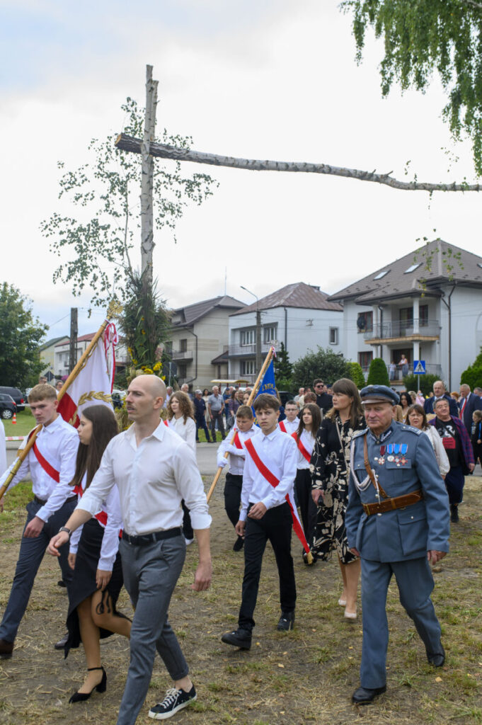 Grupa osób w białych koszulach maszeruje z flagami. W tle widać budynki mieszkalne i drzewo. Starszy mężczyzna w mundurze prowadzi pochód.