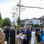 Grupa ludzi ubranych elegancko idzie po trawiastej ścieżce. Kobieta i mężczyzna niosą chleb na białych serwetach. W tle domy i krzyż z brzozy.