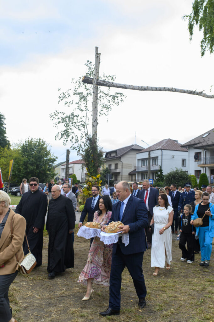 Grupa ludzi ubranych elegancko idzie po trawiastej ścieżce. Kobieta i mężczyzna niosą chleb na białych serwetach. W tle domy i krzyż z brzozy.