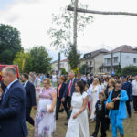 Grupa ludzi w kolorowych strojach idzie w procesji. Tło to domy i drzewa przy pochmurnym niebie. Centralnie widoczny duży krzyż.