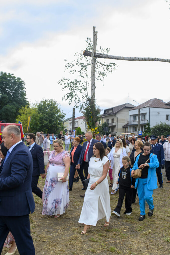 Grupa ludzi w kolorowych strojach idzie w procesji. Tło to domy i drzewa przy pochmurnym niebie. Centralnie widoczny duży krzyż.