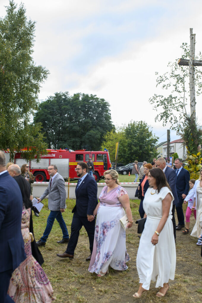 Grupa osób elegancko ubranych przechodzi obok czerwonego wozu strażackiego na tle drzew. W tle widoczny jasny niebieski niebo i zielone drzewa. Po prawej stronie znajduje się drewniany krzyż udekorowany kwiatami.