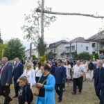 Grupa ludzi idzie w procesji przez miasto. W tle są domy i ozdobne drzewo. Uczestnicy są elegancko ubrani.