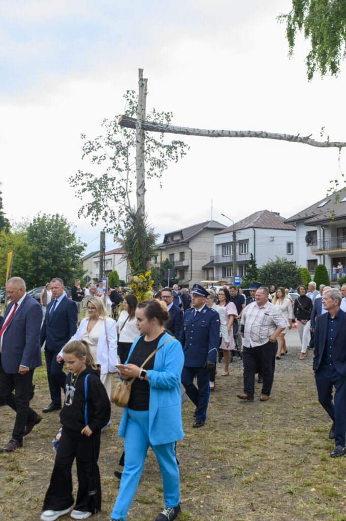 Grupa ludzi idzie w procesji przez miasto. W tle są domy i ozdobne drzewo. Uczestnicy są elegancko ubrani.